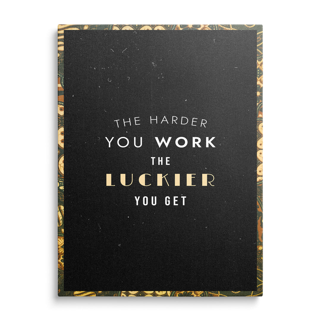 Bílý a krémový nápis The Harder You Work The Luckier You Get se zlatým slovem Luckier na černém podkladu s ornamentálním rámem.