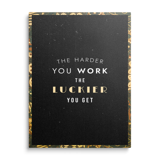Bílý a krémový nápis The Harder You Work The Luckier You Get se zlatým slovem Luckier na černém podkladu s ornamentálním rámem.