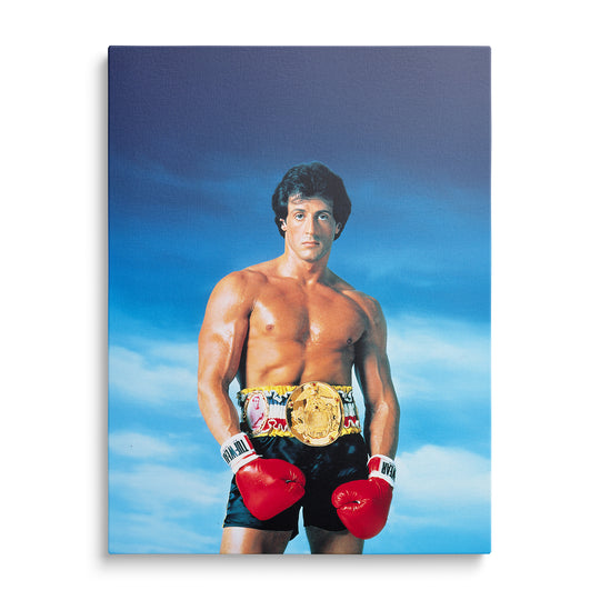 Sylvester Stallone jako Rocky v černých šortkách se zlatým šampionským pásem a červenými rukavicemi před modrou oblohou.